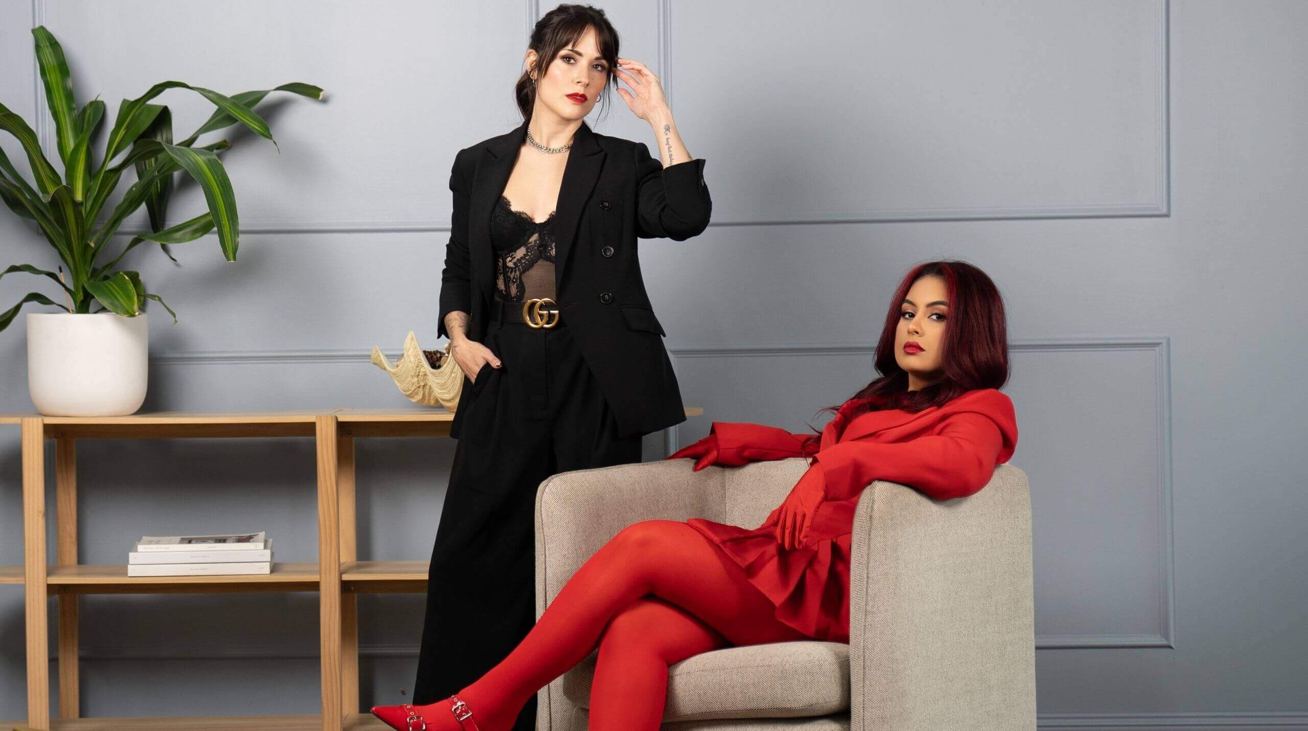 Two stylish women posing in a modern office setting, one seated in a beige armchair wearing a bold red outfit, and the other standing in a black suit with a lace top.