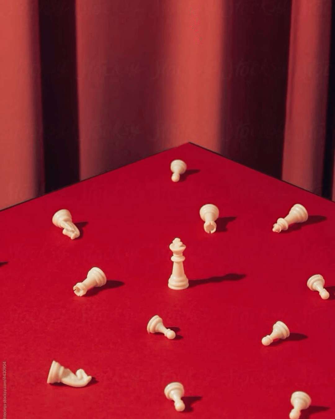 White chess queen standing upright among fallen white chess pieces on a red surface with a red curtain backdrop.