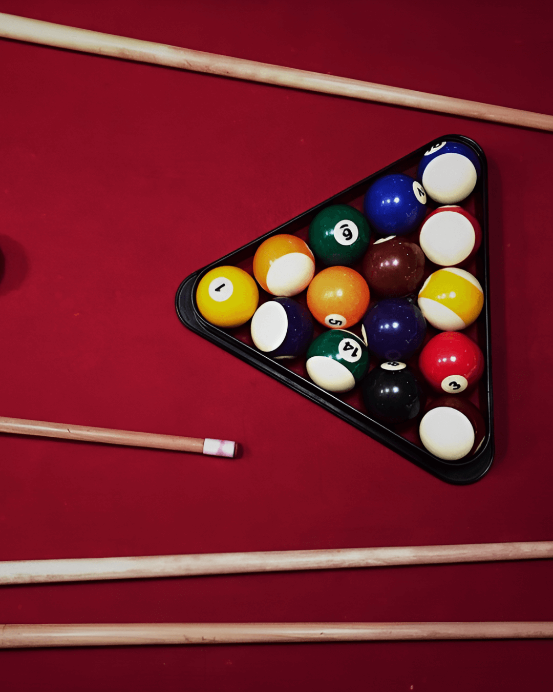 Making a bold break with red. Triangle rack of colourful billiard balls on a red pool table with wooden cues placed around it.